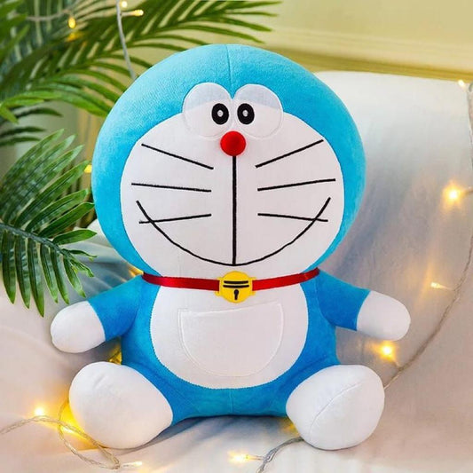 Cute blue and white Doraemon soft plush toy with red collar and yellow bell sitting against a decorated background.