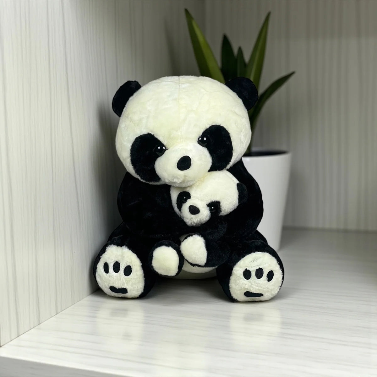 A soft, cute mother and baby panda plush toy sitting together against a light background with a decorative plant in a white pot.